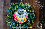 A wreath made of various green plants and colorful flowers, with a circular sign in the center that says "It's always happy hour at our house".