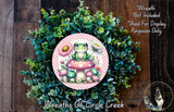 a wreath with a frog sitting on a mushroom, surrounded by various flowers and greenery. The wreath is placed on a wooden surface, and there is a small dog visible in the bottom right corner of the image.