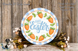 A decorative Easter wreath with the text "Happy Easter" and illustrations of carrots, placed on a wooden surface with a floral arrangement and some flowers.