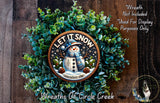 A wreath made of green leaves and a snowman decoration, with the text "LET IT SNOW somewhere else" displayed on it.