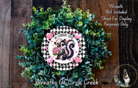 A wreath made of green and white flowers, with a black and white checkered border and a cartoon image of a skunk holding flowers.
