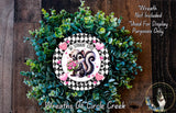 A wreath made of green and white flowers, with a black and white checkered border and a cartoon image of a skunk holding flowers.