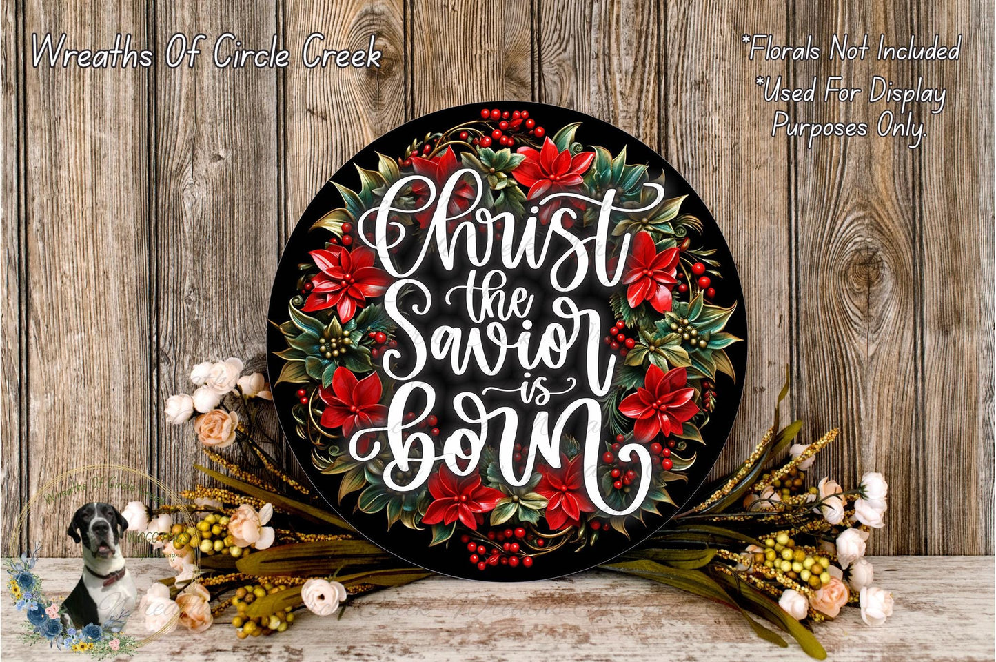 A decorative wreath with the text "Christ the Savior is Born" written on it, surrounded by red poinsettia flowers and greenery. The wreath is placed on a wooden surface, and there are some flowers and greenery visible in the foreground.