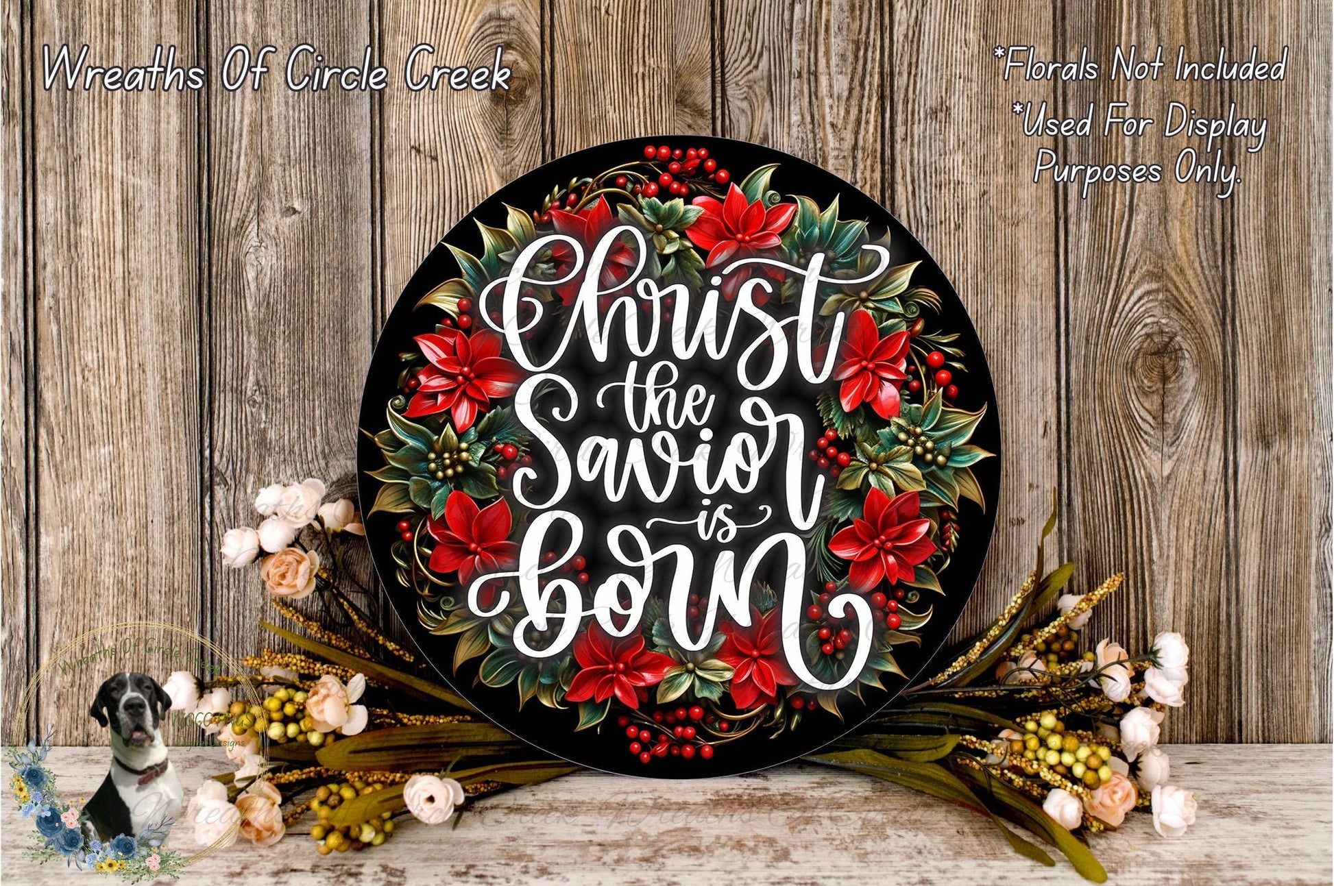 A decorative wreath with the text "Christ the Savior is Born" written on it, surrounded by red poinsettia flowers and greenery. The wreath is placed on a wooden surface, and there are some flowers and greenery visible in the foreground.