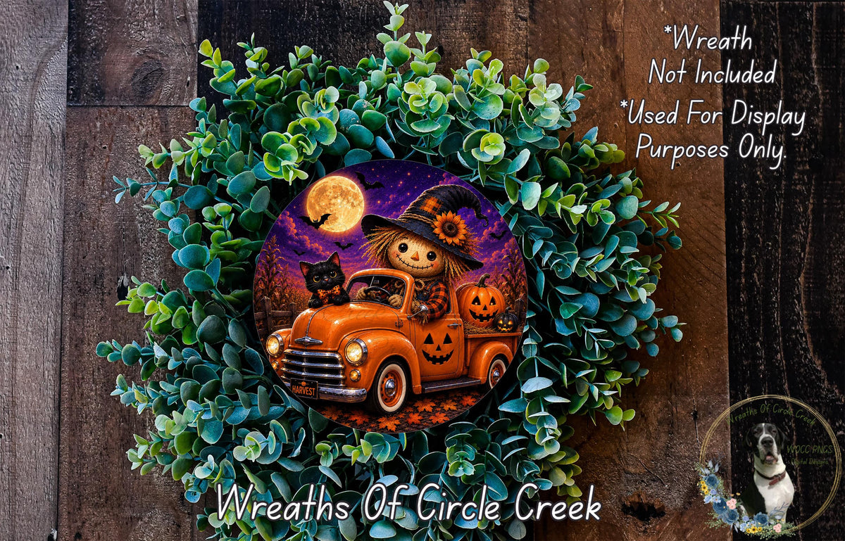 A Halloween-themed wreath featuring a scarecrow in a truck, a jack-o-lantern, and a full moon. The wreath is surrounded by greenery and placed on a wooden surface.