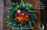 A Halloween-themed wreath featuring a scarecrow in a truck, a jack-o-lantern, and a full moon. The wreath is surrounded by greenery and placed on a wooden surface.