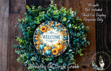 a wreath made of various green and orange foliage, with a central circular design that says "WELCOME". The wreath is displayed on a wooden surface, and there is a small dog in the bottom right corner of the image.