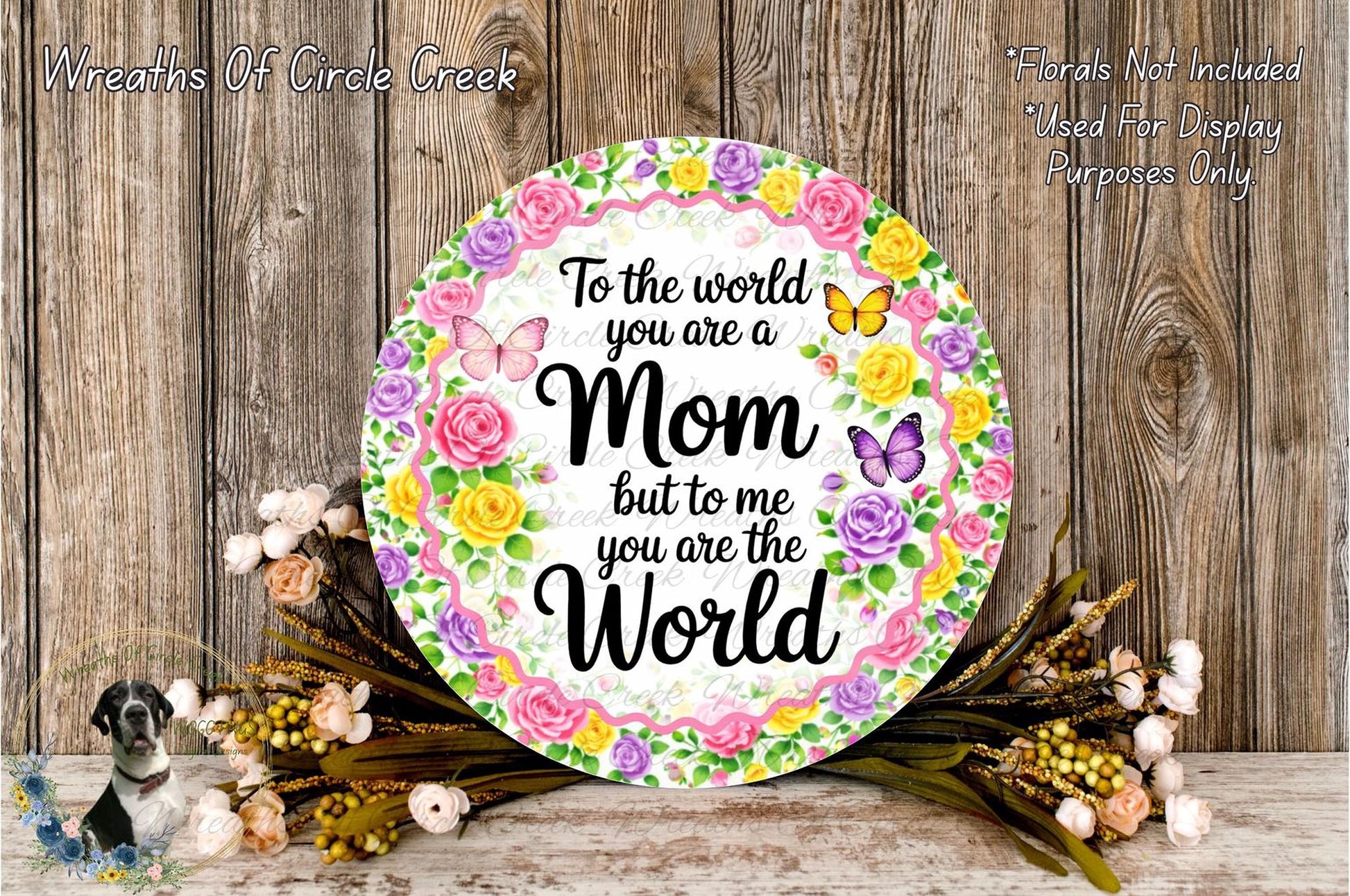a round floral wreath with a message that reads "To the world you are a Mom but to me you are the World". The wreath is surrounded by various flowers and placed on a wooden surface.
