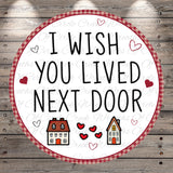a circular design with the text "I WISH YOU LIVED NEXT DOOR" written in a playful font. The design is surrounded by small illustrations of houses and hearts, creating a whimsical and romantic atmosphere.