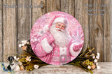 A round, decorative wreath featuring a smiling Santa Claus figure wearing a pink outfit, surrounded by a floral arrangement and placed on a wooden surface.