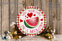 A round wooden sign with a watermelon slice and hearts, surrounded by flowers and placed on a wooden surface.