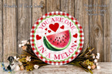 A round wooden sign with a watermelon slice and hearts, surrounded by flowers and placed on a wooden surface.