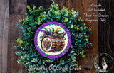 a wreath with a decorative drum design at the center, surrounded by a lush green plant. The wreath is placed on a wooden surface, and there is a small dog visible in the bottom right corner of the image.
