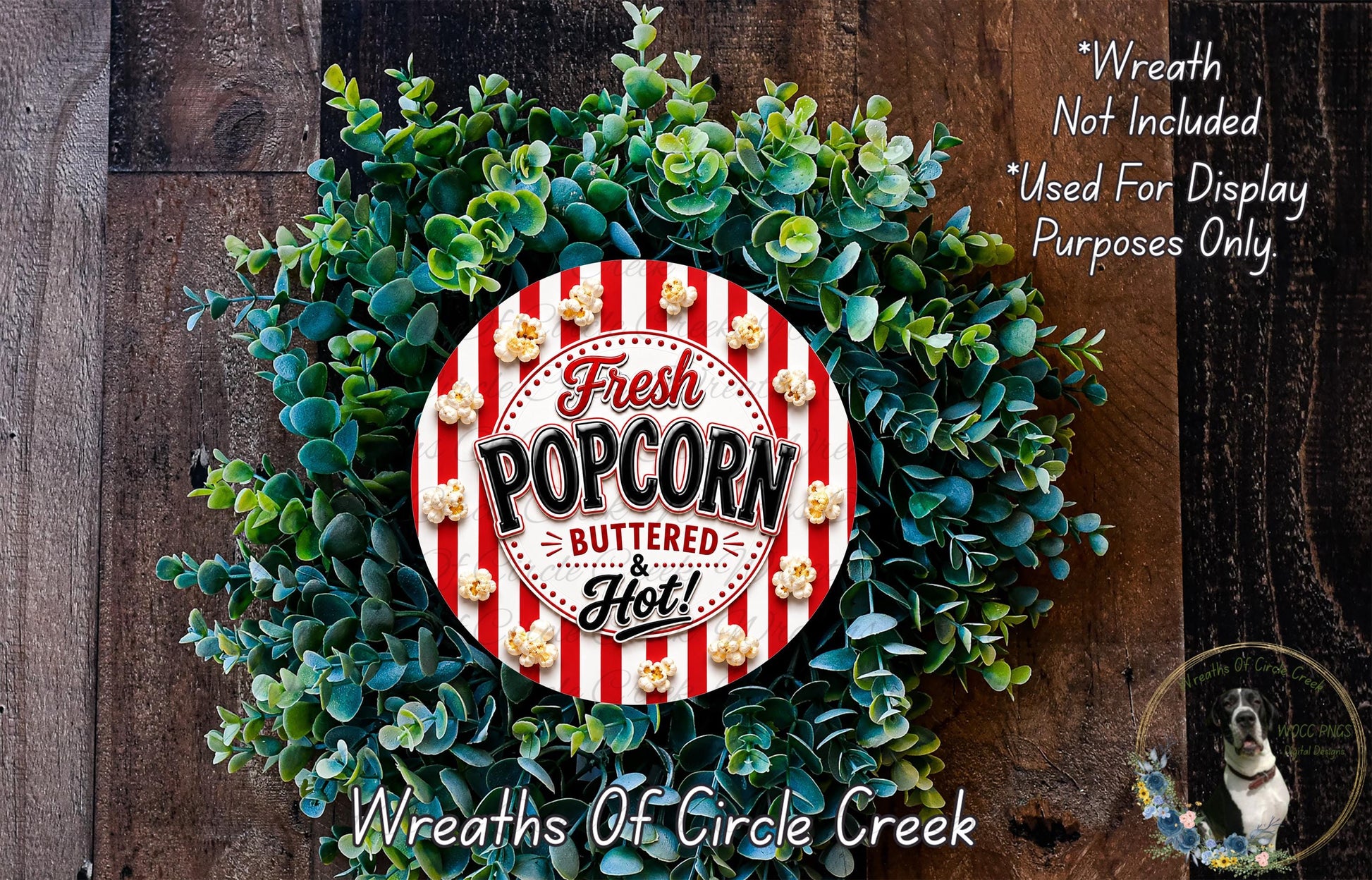 a wreath made of green leaves and a red and white striped circular sign that says "Fresh Popcorn Buttered & Hot!". The sign is surrounded by the wreath, which is placed on a wooden surface.