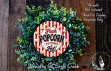 a wreath made of green leaves and a red and white striped circular sign that says "Fresh Popcorn Buttered & Hot!". The sign is surrounded by the wreath, which is placed on a wooden surface.
