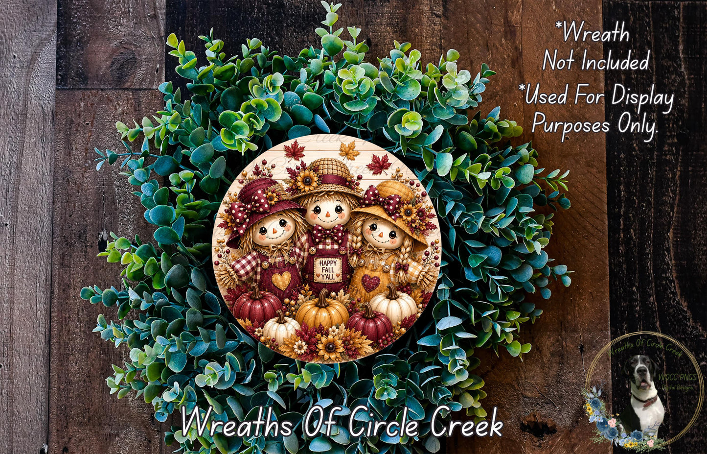 a wreath with three scarecrow dolls and pumpkins, surrounded by a lush green plant. The wreath is displayed on a wooden surface, and there is a small dog in the bottom right corner of the image.