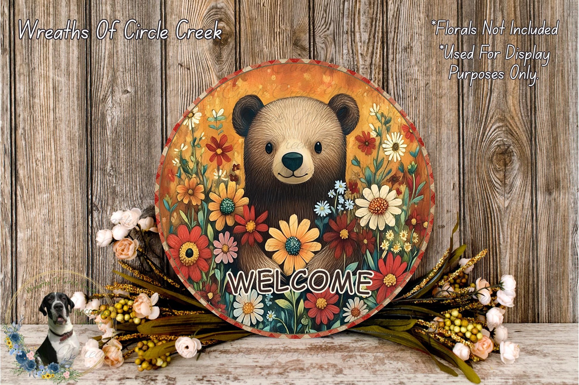 A round sign with a bear surrounded by a colorful floral arrangement, with the word "WELCOME" written on it. The sign is placed on a wooden surface, and there are some flowers and greenery around it.