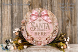 A decorative sign with the text "Santa stops here" surrounded by floral elements, including a bow and various ornaments, placed on a wooden surface.