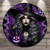 A woman with long, dark hair and makeup, wearing a black hat and holding a carved pumpkin lantern, set against a purple and black background with a wooden wall.