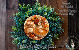 A wreath made of green leaves and flowers, with a pumpkin pie and other fall-themed items displayed on it.