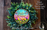 A colorful wreath made of various plants and flowers, with the text "Hello Spring" displayed on it. The wreath is placed on a wooden surface, and there is a small dog in the bottom right corner of the image.