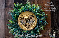 a wreath made of green plants and a heart-shaped ornament with the text "Be Mine" on it. The wreath is displayed on a wooden surface, and there is a small dog in the bottom right corner of the image.