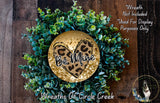 a wreath made of green plants and a heart-shaped ornament with the text "Be Mine" on it. The wreath is displayed on a wooden surface, and there is a small dog in the bottom right corner of the image.