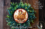 a wreath with a large "GOBBLE" sign in the center, surrounded by a lush green wreath. The wreath is displayed on a wooden surface, and there is a small dog visible in the bottom right corner of the image.