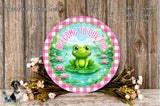 A round sign with a green frog sitting on a lily pad, surrounded by pink flowers and a wooden background.