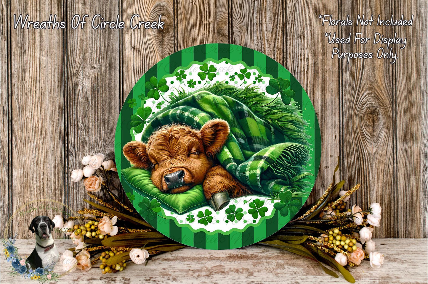 A circular green and white wreath with a sleeping cow wrapped in a green blanket, surrounded by flowers and a wooden background.