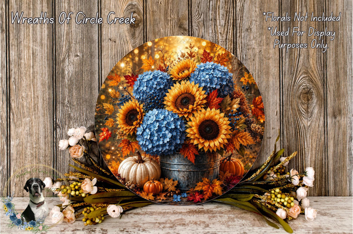 A round, wooden-paneled display featuring a vibrant arrangement of blue hydrangeas, sunflowers, and pumpkins, with additional fall foliage and flowers.