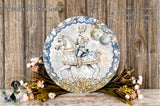 A decorative plate featuring a man in a blue and gold outfit riding a white horse, surrounded by a wreath of flowers and greenery.
