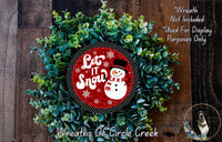 A wreath made of green plants and a red circular sign with a snowman and the text "Let It Snow" is displayed on a wooden surface.