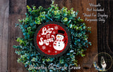 A wreath made of green plants and a red circular sign with a snowman and the text "Let It Snow" is displayed on a wooden surface.
