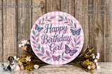 A round sign with the text "Happy Birthday Girl" surrounded by a wreath of flowers and butterflies, placed on a wooden surface.