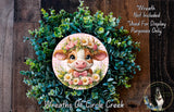 a wreath with a cow's face and floral elements, including a pink bow and flowers, set against a backdrop of greenery.