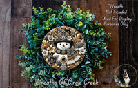 A decorative wreath made of various green plants and a snowman figure, with a wooden background.