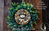 A decorative wreath made of various green plants and a snowman figure, with a wooden background.