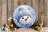 A snowman decoration with a blue scarf and hat, surrounded by flowers and placed on a wooden surface.