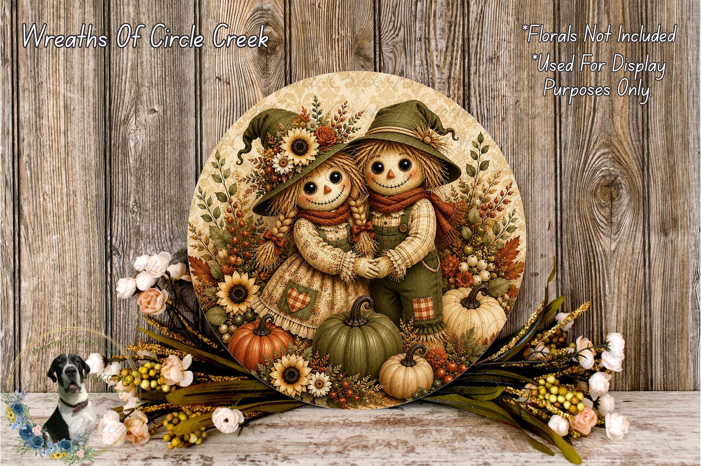 A decorative wreath featuring two scarecrow figures, surrounded by autumn foliage, pumpkins, and flowers, placed on a wooden surface.
