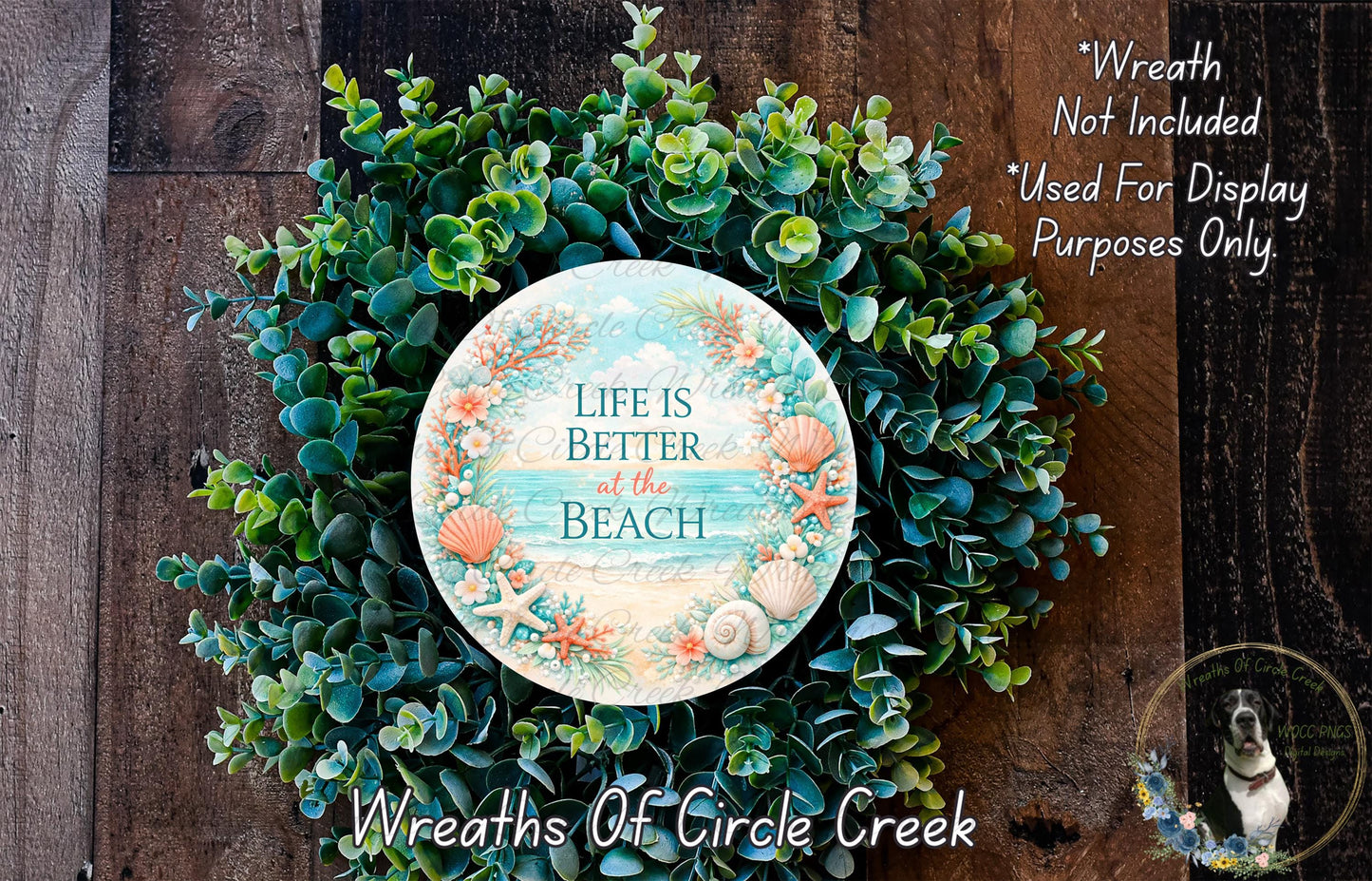 a wreath made of various green plants and a circular sign with the text "Life is better at the beach" on it. The wreath is displayed on a wooden surface, and there is a small dog in the bottom right corner of the image.
