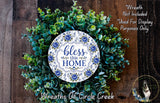 a wreath made of various green plants and flowers, with a decorative plate in the center that says "bless this home".