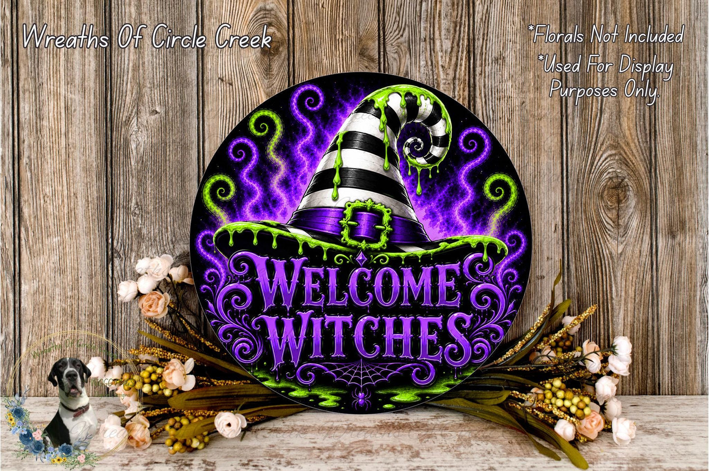 a decorative sign with a witch's hat and the text "Welcome Witches" against a purple and green background. The sign is surrounded by flowers and placed on a wooden surface.