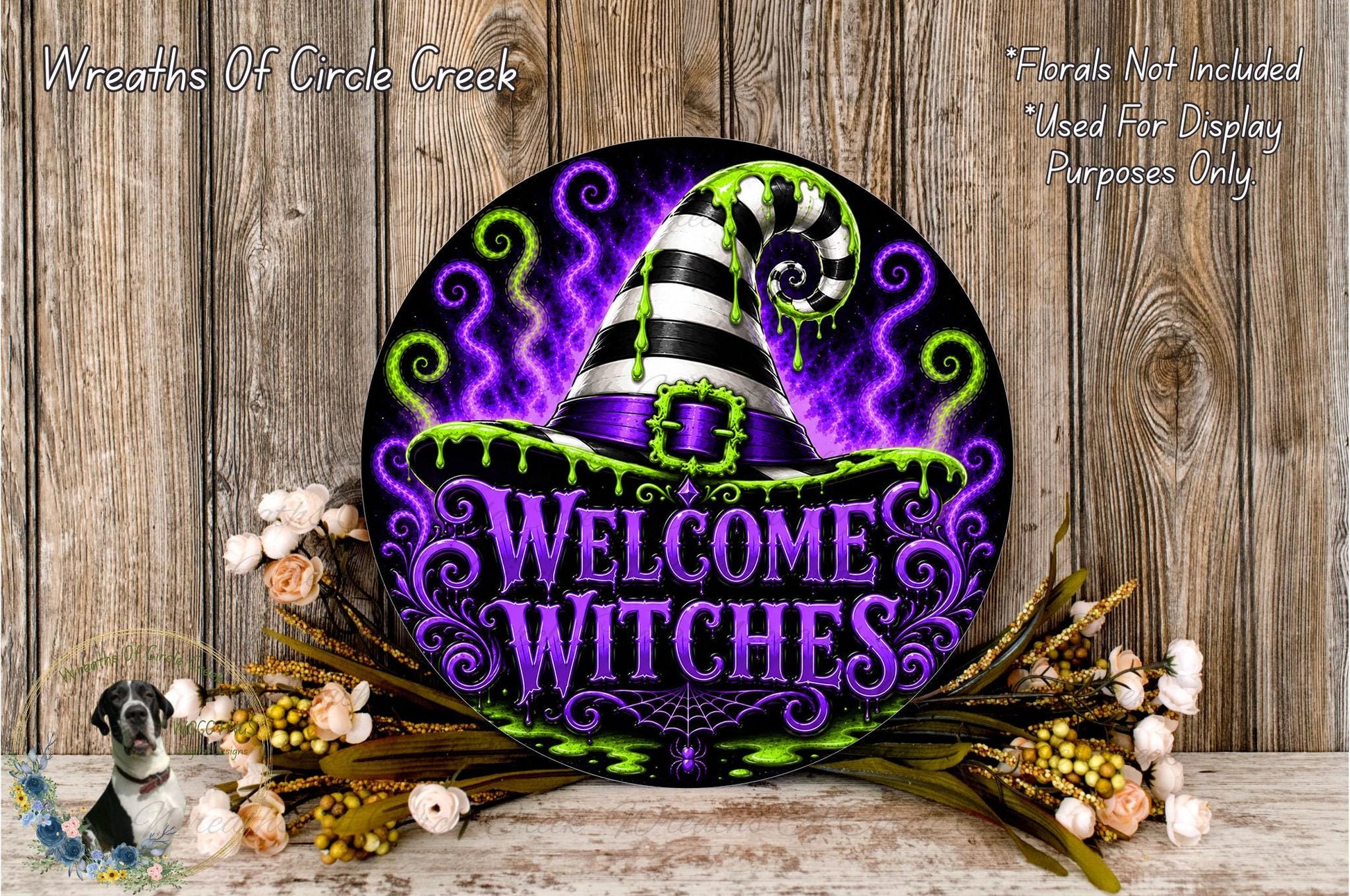 a decorative sign with a witch's hat and the text "Welcome Witches" against a purple and green background. The sign is surrounded by flowers and placed on a wooden surface.