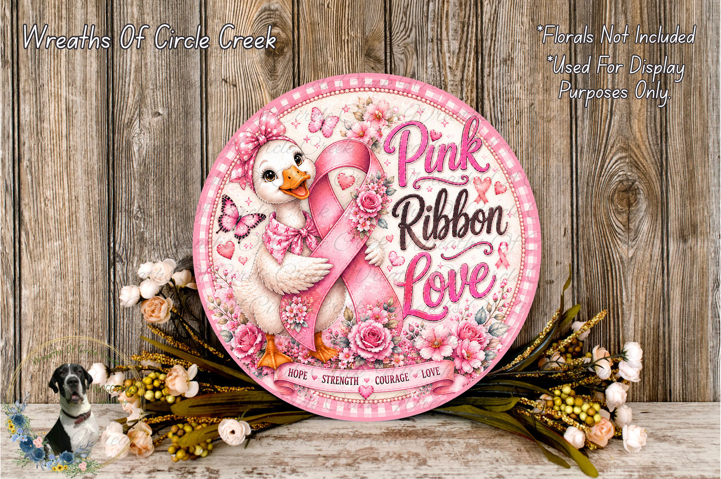 A decorative sign featuring a pink ribbon and a duck holding a pink ribbon, surrounded by flowers and placed on a wooden surface.