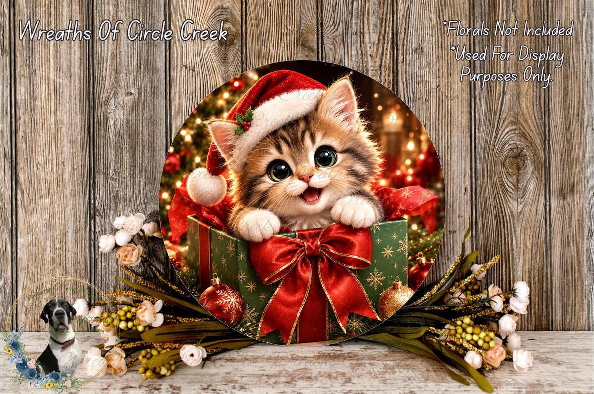 A cute kitten wearing a Santa hat is sitting in a gift box surrounded by a wreath of flowers and ornaments, with a wooden background.