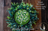 A wreath made of green leaves and a large four-leaf clover, adorned with sparkling jewels, is displayed on a wooden surface.