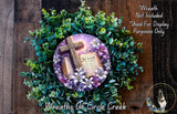 A wreath made of green and purple flowers, with a wooden cross and a book titled &quot;Jesus&quot; displayed on it.