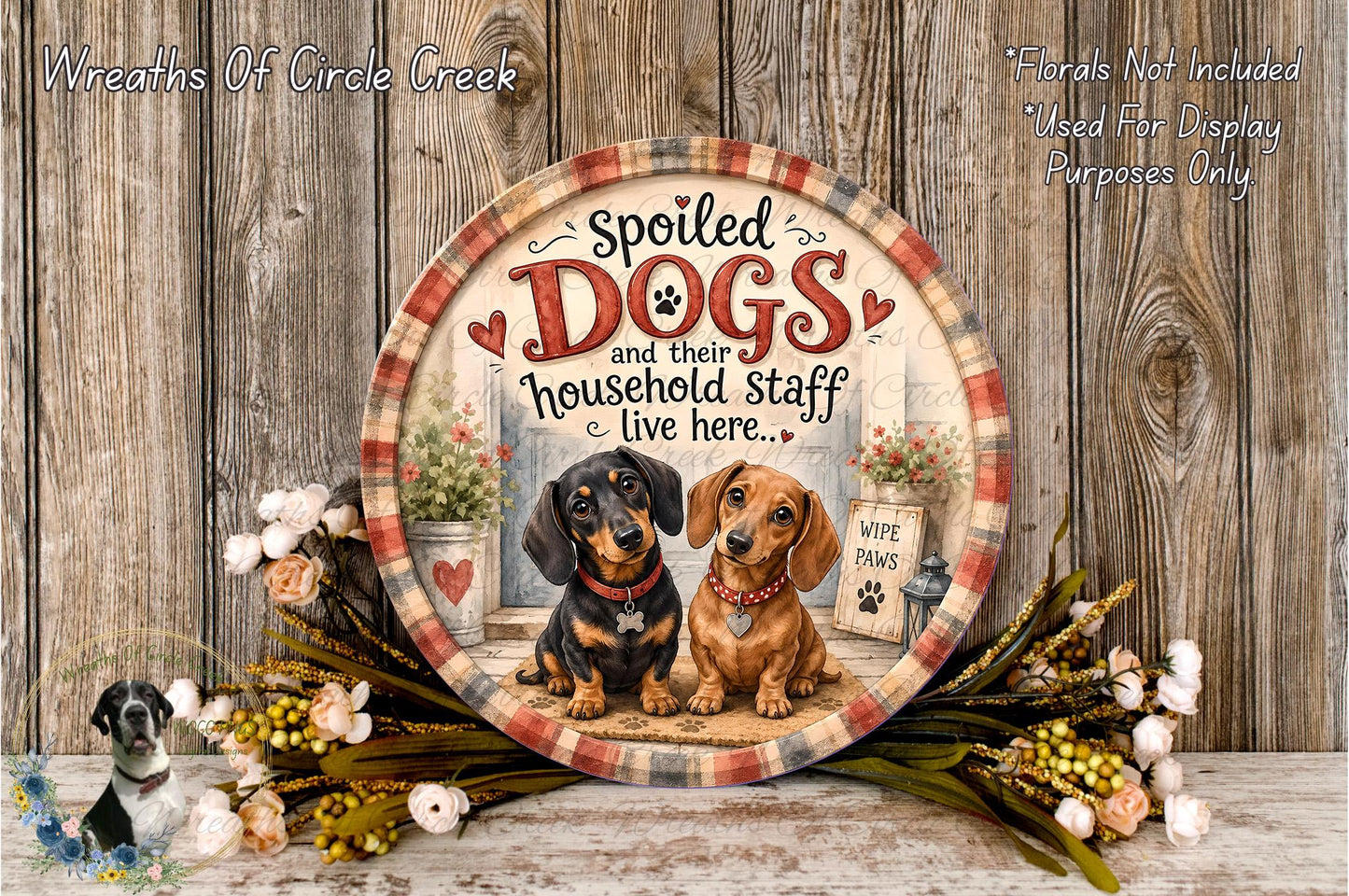 a decorative plate with a painting of two dogs, one black and one brown, sitting together in front of a house. The plate is surrounded by a wreath of flowers and a wooden wall.