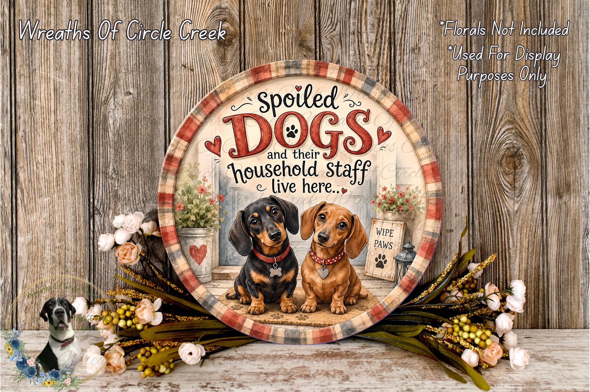 a decorative plate with a painting of two dogs, one black and one brown, sitting together in front of a house. The plate is surrounded by a wreath of flowers and a wooden wall.